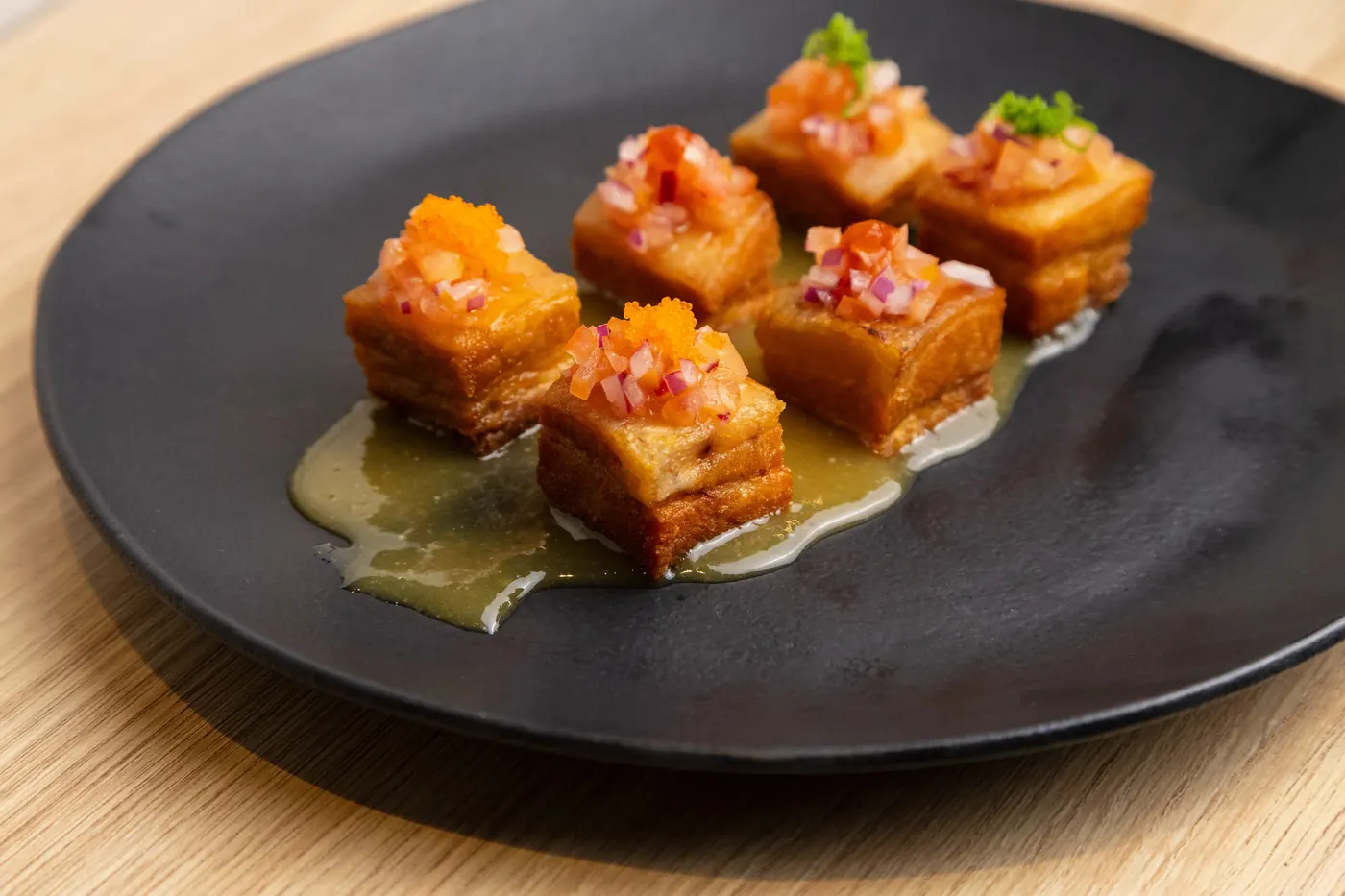 O Norū Pork: seis cubos dourados de porco, com ovas laranja e cebola roxa picada por cima, sobre um molho claro, num prato preto.