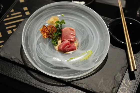 Sashimi de atum com uma flor e salsa em prato de vidro, sobre ardósia, com hashis dourados ao lado.