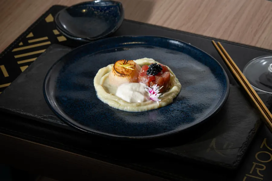 Vieira maçaricada e tartare de atum com ovas pretas sobre purê, em prato azul-escuro, com hashis dourados ao lado.